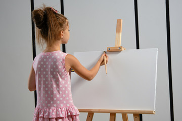 Wall Mural - Little girl artist in a pink dress is standing behind easel and painting with brush on canvas at art studio with white walls. Medium close-up shot.