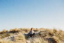 Low Angle View Of Boy Sitting On Sand Dune Against Clear Blue Sky