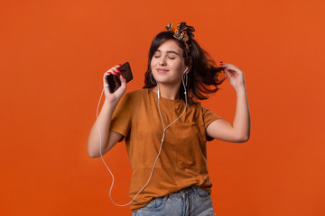 Pretty brunette woman in a t-shirt and beautiful headband dancing listening to music with wired headphones isolated over orange background. Enjoying life