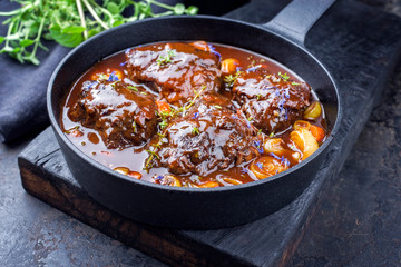 Traditional German braised beef cheeks in brown red wine sauce with carrots and onions offered as closeup in a cast iron Dutch oven on an old rustic cutting board