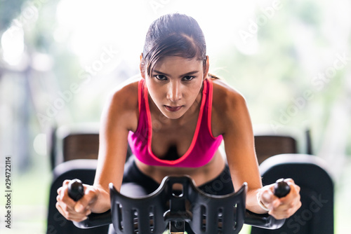 Fototapeta na szkło Piękna Azjatycka kobieta ćwiczy na stacjonarnej kolarstwo maszynie w salowym sprawności fizycznej gym, determinaci twarz. Aktywność sportowa, trening ludzi lub pojęcie zdrowego stylu życia