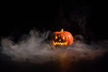 Halloween, Orange Pumpkin With A Scary Luminous Face On A Dark Background. Thick Gray Smoke Comes Out And Spreads Across The Black Table. A Close-up Of A Flashlight On The Eve Of All The Saints