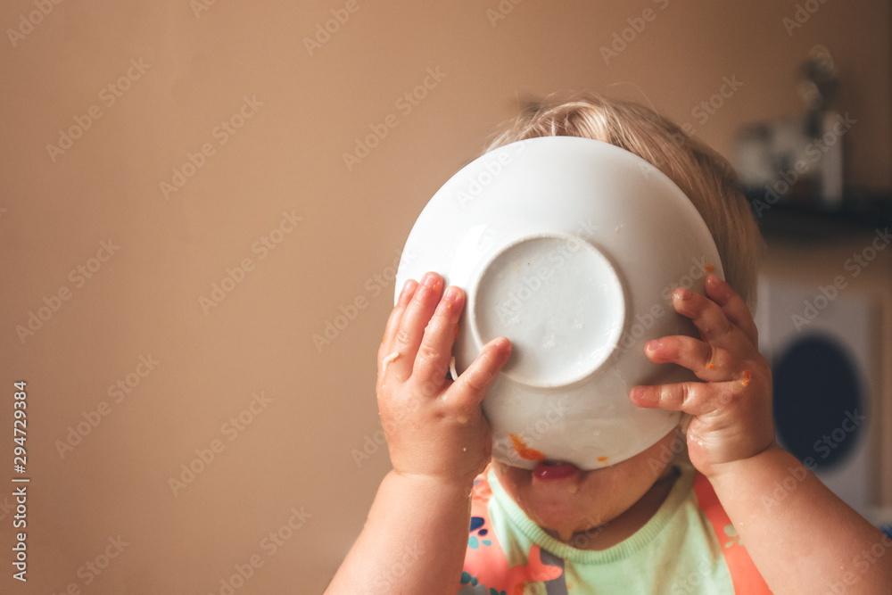 blond kid boy, very hungry eats and holds a plate with both hands, close-up with copyspace - obrazy, fototapety, plakaty 