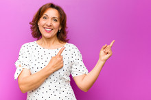 Middle Age Woman Smiling Happily And Pointing To Side And Upwards With Both Hands Showing Object In Copy Space Against Purple Wall