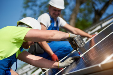 Close-up of two professional technicians connecting solar photo voltaic panel to metal platform on blue sky background. Stand-alone solar system installation, efficiency and professionalism concept.