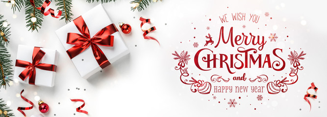 Wall Mural - Merry Christmas text on white background with gift boxes, fir branches, red decoration. Xmas and New Year greeting card, bokeh, light. Flat lay, top view, winter holiday, wide compositon