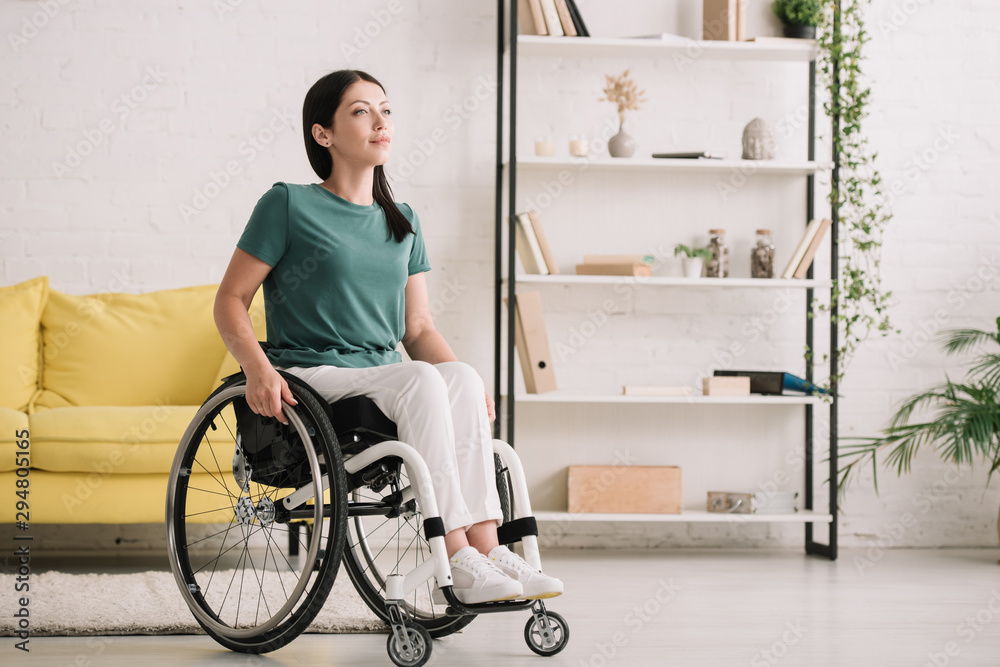 smiling disabled woman looking away while sitting in wheelchair at home - obrazy, fototapety, plakaty 