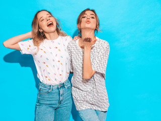 Two young beautiful smiling blond hipster girls in trendy summer colorful T-shirt clothes. Sexy carefree women posing near blue wall. Positive models having fun and shows funny grimace