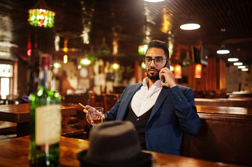 Wall Mural - Handsome well-dressed arabian man with glass of whiskey and cigar hold mobile phone,  posed at pub.