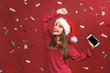 © denis_vermenko - Cheerful attractive young woman in santa claus hat dancing and having fun isolated over red background. Phone screen.