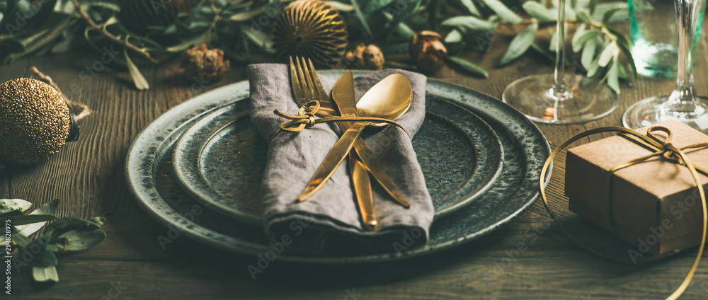 Christmas or New Years celebration party table setting. Plates, golden cutlery, glasses, gift box, festive branch decoration, candles, gliterring toys over rustic wooden table background - obrazy, fototapety, plakaty 