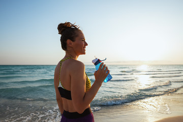 young woman with bottle of water on ocean coast at sunset