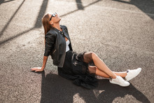Joyful Young Hipster Woman In Fashionable Jacket In A Skirt In A Sneakers In Stylish Sunglasses Sits On The Asphalt On A Sunny Autumn Day. Positive Girl Model With A Cute Smile Is Resting On A Sun.
