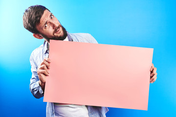 Wall Mural - Handsome bearded caucasian man holding blank pink poster board in hands with copy space for text or advertising inscription isolated in blue studio