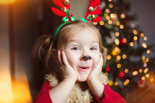 Cute Funny Baby Is Very Surprised. Pretty Girl In A Fancy Dress Deer On The Background Of The Christmas Tree At Home. Shocked And Humorous Kid