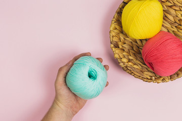 Human hand holding a blue ball of wool near to a basket of pink and yellow balls of wool on a pink background