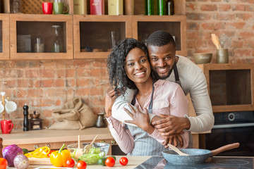 Wall Mural - Cheerful black man hugging his wife while cooking at kitchen