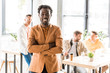 © LIGHTFIELD STUDIOS - cheerful african american businessman standing with crossed arms near colleagues in office and smiling at camera