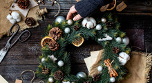Overhead Shot Of Handmade Christmas Wreath With Grey Balls, Pine Cones, Dried Orange Slices, Cinnamon Sticks In Woman Hand On Rustic Wooden Table With Old Scissors, Sackcloth, New Year Toys