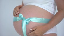 Close Up Of Woman Pregnant Belly With Blue Ribbon Bow. Mom Anticipation Expecting Baby, Pregnant Woman Touching Belly On White Background. Newborn Boy. Pregnancy Maternity Motherhood, Mother Hands 4 K