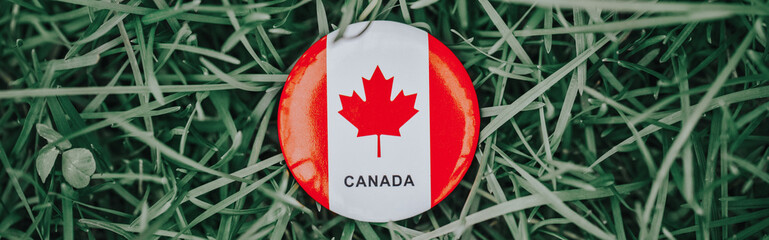 Round circle badge with red white canadian flag maple leaf lying in grass on green forest nature background. Web banner header for website. Canada day holiday national celebration.