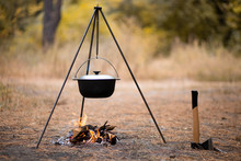 Campfire With Camping Tools Such As Axe And Hanging Pot Over Fire. Secluded Relaxation In Autumn Forest Away From Bustle Of City