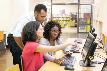 Wall Mural - instructor helping students in computer class. man and women sitting and standing at desk, using des