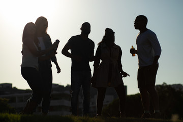 Group of friends having fun at outdoor party. Silhouettes of multiethnic men and women drinking beer and dancing on grass. Relaxing outdoors concept