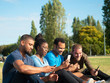 Multiethnic friends sharing content on phones while relaxing on park lawn. Young men and woman sitting on grass, using smartphones, smiling, laughing. Communication concept