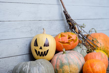 Pumpkins And Jack Lantern Of Different Colors And Sizes Lie On Gray Wooden Wall Background With Jug And Pumpkins Hanging On It. Concept Of Halloween Celebration And Joy Of Harvest Period. Copyspace