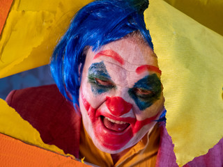 Portrait of male clown in pink dressed for Halloween. actor made makeup from crazy evil clown, faced with bloody smile. White face makeup. Colour wig. Model in Halloween costume rips yellow paper