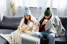 Attractive Girlfriend Reading Book And Boyfriend In Winter Outfit With Cup Warming Up Near Heater