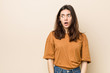 © Asier - Young brunette woman against a beige background being shocked because of something she has seen.
