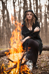 Young woman in black cape sitting near bonfire.