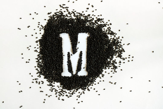Handful of sweet basil black seeds, letter M uppercase of English alphabet on white background. Black and white. Lettering. Typography. Healthy and super food. Horizontal