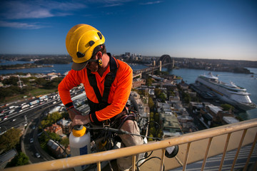 Wall Mural - Rope access industrial worker wearing yellow hard hat, long sleeve shirt, safety harness, inspecting safety white bucket prior to abseiling down from the high rise building at circular quay, Sydney