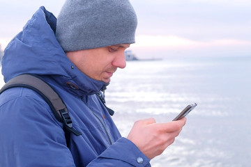 Portrait of man is typing message on smartphone in coastal town at sea in winter, side view. Man traveller freelancer blogger is writing post on phone. Tourist travel journey trip with backpack.