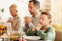 Little Girl Sitting At The Table With Eyes Closed And Praying Together With Her Family During Holiday Dinner