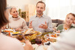 © AnnaStills - Young man sitting at dining table and talking to the members of his family during family dinner at home