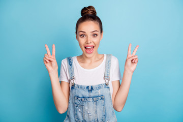 Wall Mural - Portrait of cheerful girl screaming making v-signs wearing white t-shirt denim jeans overall isolated over blue background