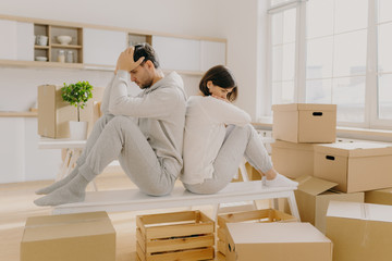 Unhappy young married woman and man have to leave house, move in other place, sit back to each other pose in empty room with stack of boxes, wear domestic clothes and socks, have some problems