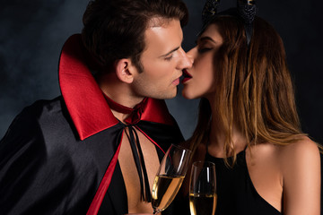 handsome man in cloak and woman with horns kissing and holding champagne glasses on black with smoke