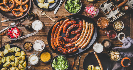 Wall Mural - Flat-lay of Octoberfest dinner table concept with grilled veal and pork sausages, pretzel pastry, potatoes, cucumber salad, sauces, beers in bottles and glasses over dark wooden background, top view