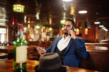 Wall Mural - Handsome well-dressed arabian man with glass of whiskey and cigar hold mobile phone,  posed at pub.