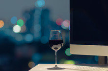 A Glass Of Red Wine Put On Working Table, Computer Screen And Keyboard With Bokeh Of City Lights.