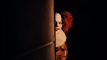 Creepy Clown With Crazy Smile On His Face Suddenly Appears Around The Corner In Darkroom. Terrible Clown With A Colorful Makeup In A Festive Costume. Shooting In Slow Motion