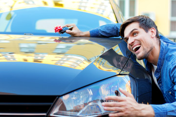 Happy cheerful owner of new black car is holding gift, surprise, keys with red bow. Smiling brunette driver in denim shirt is hugging and stroking automobile. Man winner won auto in raffle.