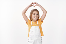Lovely Tender Friendly Young Girl With Blond Hair, Wear Summer Yellow T-shirt, Overalls, Raise Hands Up Show Heart Sign, Confess Love To Parents, Cherish Friendship, Smiling Joyful, White Background
