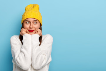 Wall Mural - Horizontal shot of happy dreamy European woman wears minimal makeup, red lipstick, looks aside, dressed in yellow hat and white sweater, poses against blue background, being fascinated and pleased