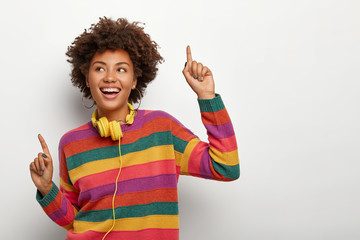 Wall Mural - Cheerful Afro American woman raises arms and points with index fingers, dances happily to music, wears striped colored sweater and stereo headphones, has overjoyed expression, models indoor.